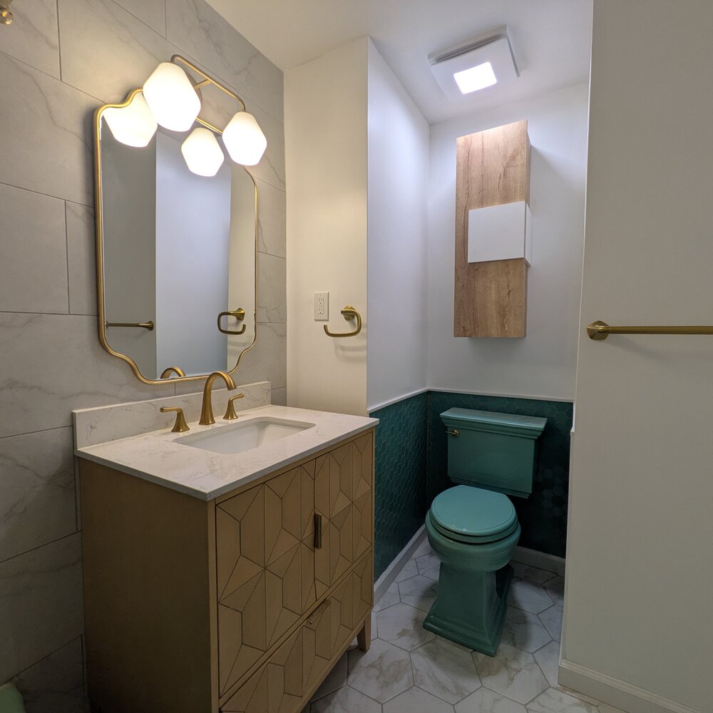 Bathroom with teal toilet, green hexagon tile wainscot, patterned wood vanity, brass mirror and fixtures, and marble-look tile