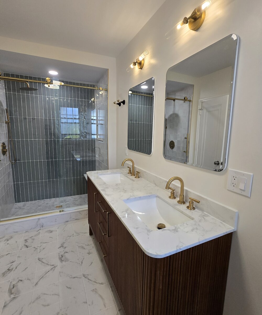 Bathroom with fluted walnut double vanity, brass fixtures, and sea-green tiled walk-in shower with brass sliding glass door